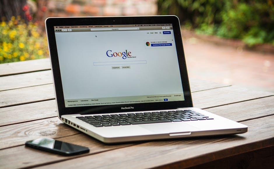一台 MacBook Pro 放在户外的木质桌子上，屏幕显示 Google 搜索，旁边放着一部智能手机